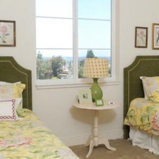 A cozy bedroom with twin beds featuring green headboards and floral bedding, separated by a side table with a lamp under a window.