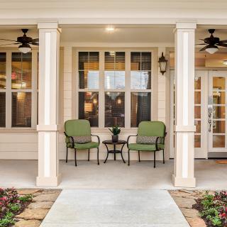 A welcoming porch with green chairs and a small table, framed by columns and illuminated by ceiling fans and outdoor lighting.
