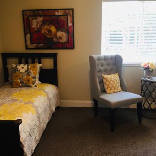 A cozy bedroom features a bed with floral bedding, a plush chair, and decorative elements like a painting and flowers.