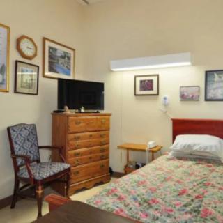 A cozy bedroom featuring a bed with floral bedding, a wooden chest of drawers topped with a small television, and walls adorned with various framed pictures.