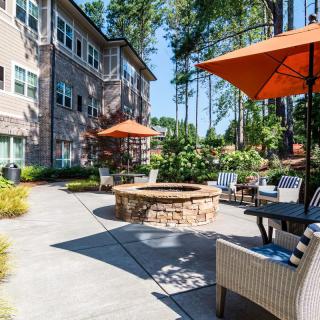 A cozy outdoor patio area with a fire pit and seating at an apartment complex, surrounded by lush landscaping and trees.