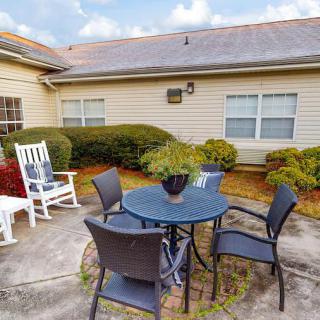 Outdoor patio area with a table, chairs, and a rocking chair surrounded by shrubs.