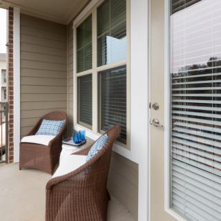 A cozy balcony with wicker chairs and cushions, next to a door and windows with blinds.