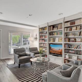 A modern living room featuring built-in bookshelves, comfortable armchairs, and a striped rug, with ample natural light streaming in through large windows.