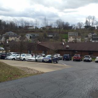A view of a parking lot with various vehicles, surrounded by buildings and a hilly landscape under a cloudy sky.