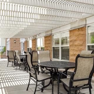 A patio area with tables and chairs under a pergola, casting shadows in the sunlight.