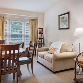 A cozy living room with a dining area, featuring a beige sofa, wooden dining table, and natural light from the window.