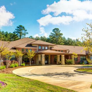 A beautiful residential building with a wide driveway, surrounded by trees and a sunny, cloudy sky.