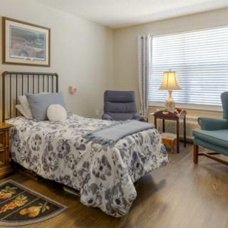 A cozy bedroom featuring a bed with patterned bedding, a comfortable chair, and a lamp by the window.