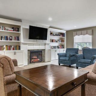 A cozy living room with a fireplace, surrounded by bookshelves, armchairs, and a television above.