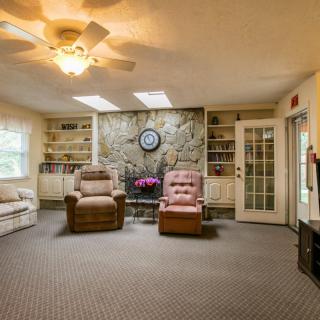A cozy living room featuring comfortable armchairs, a soft carpet, ceiling fan, skylights, and built-in bookshelves surrounding a central stone fireplace.