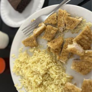 A plate of noodles and breaded chicken with a slice of cake in the background.