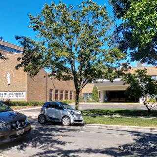A sunny day in an urban setting featuring parked cars and a tree beside a building.