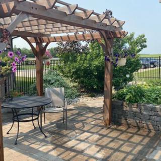 A cozy garden patio with a wooden pergola and outdoor seating, surrounded by lush greenery and flowers.