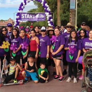 A group of people participating in an outdoor charity walk, wearing purple shirts and holding flowers, with balloons in the background.