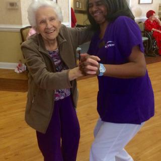 An elderly woman joyfully dances with a caregiver in a nursing home setting.