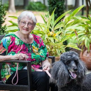 An elderly woman sitting outdoors in a garden with her poodle by her side.