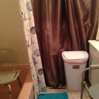 A small bathroom with a shower curtain, trash can, and chair.