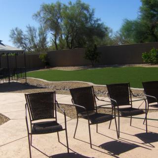 A sunny backyard with patio chairs arranged on a paved area, overlooking a grassy lawn enclosed by a fence.