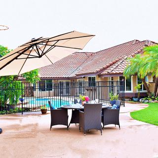 A spacious patio area with a dining set under a large umbrella, adjacent to a pool, with a house and lush greenery in the background.