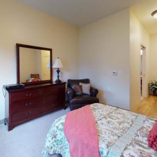 A cozy bedroom featuring a bed with a patterned bedspread, a wooden dresser with a mirror, and a comfortable chair beside a lamp.