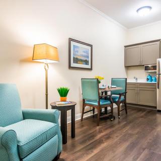 A cozy living room area with a light blue armchair, wooden flooring, and a kitchenette in the background.