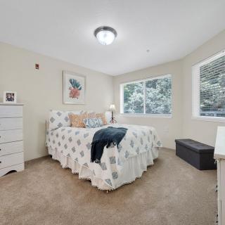 A cozy bedroom with a neatly made bed, a dresser, and large windows providing natural light.
