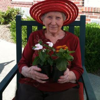 An elderly woman wearing a red hat is sitting in a rocking chair outdoors, holding a pot of flowers.