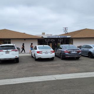 A view of a parking lot in front of a building with several cars and people walking by.