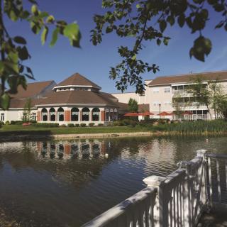 A picturesque view of a building by a pond, surrounded by trees with a white railing in the foreground.