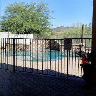 A serene view of a swimming pool surrounded by a fence, with a clear blue sky and lush greenery.
