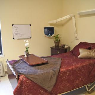 A hospital room with a neatly made bed, bedside table, monitor, and a whiteboard on the wall.