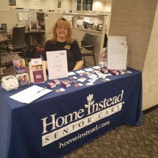 A representative sits at an informational booth for Home Instead Senior Care, showcasing promotional materials and brochures.