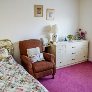 A cozy bedroom with a floral bedspread, an armchair, and a dresser on a pink carpet.