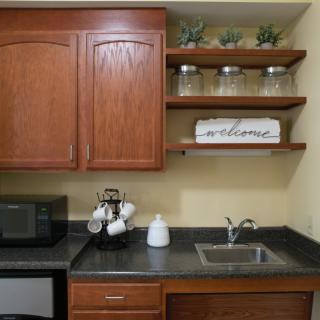 A cozy kitchen corner featuring wooden cabinets, open shelves with decorative plants and jars, a microwave, and a small sink.