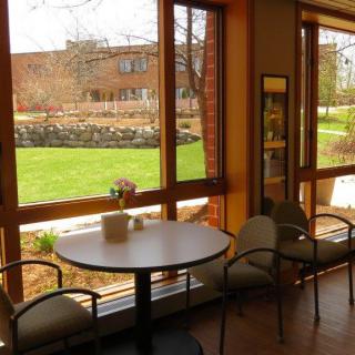 A cozy cafe interior with a round table and chairs by a large window overlooking a green outdoor area.