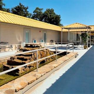 A sunny outdoor area featuring a ramp with handrails, rocks, and a building with a yellow roof.