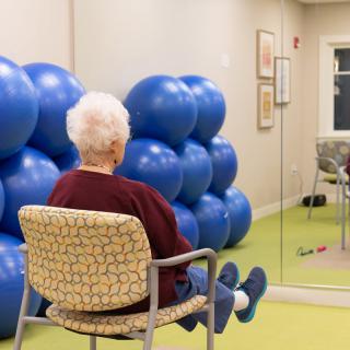 An elderly person sitting on a chair in front of a mirror, surrounded by blue exercise balls.
