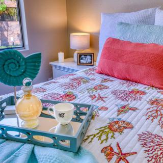 A cozy bedroom with coastal-themed bedding and decor, featuring a small lamp and a tray with a drink on the bed.