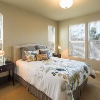A cozy bedroom with a neatly made bed, bedside tables, and large windows with blinds.