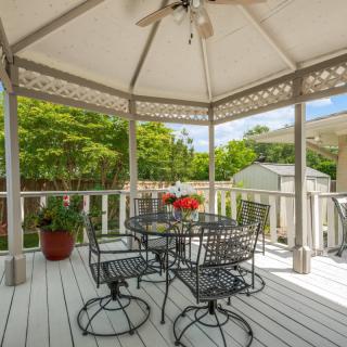 A cozy outdoor patio with a gazebo, featuring metal chairs and a table, surrounded by greenery.