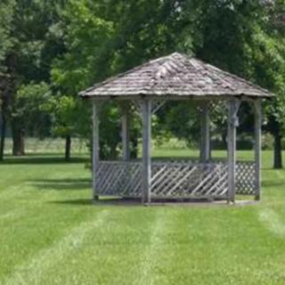 A wooden gazebo surrounded by lush green grass and trees in a serene park setting.