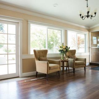 A cozy living room interior with natural light streaming through large windows, featuring two armchairs and a small table with a flower arrangement.