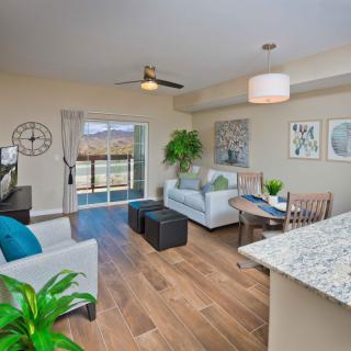 A modern and cozy living room featuring a comfortable sofa, wooden flooring, and ample natural light from large windows. The space is enhanced with indoor plants and stylish decor.