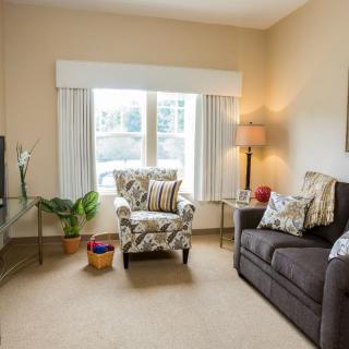 A cozy living room with a patterned armchair, a sofa, and a lamp beside a large window.