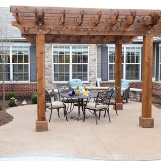 A wooden pergola over a patio with outdoor furniture and a table set, located in front of a building with multiple windows.