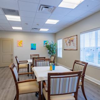 A cozy dining room featuring tables and chairs, decorated with artwork and a potted plant, illuminated by natural light from the windows.