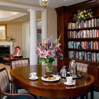 A cozy reading room with a wooden table, flowers, and a bookshelf filled with books.