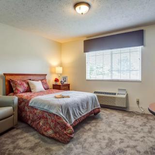 A cozy bedroom featuring a bed with floral bedding, a comfortable armchair, and a large window letting in natural light.