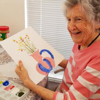 An elderly woman happily showing her painting of a vase with flowers.